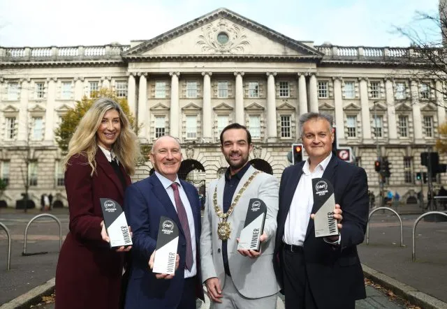 1 women and 3 men standing outside the back of city hall building, each holding an award. The awards are black and grey