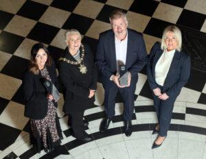 Group of 4 people standing on a black and white checked floor holding an award. One person is wearing the Lord Mayor Chain