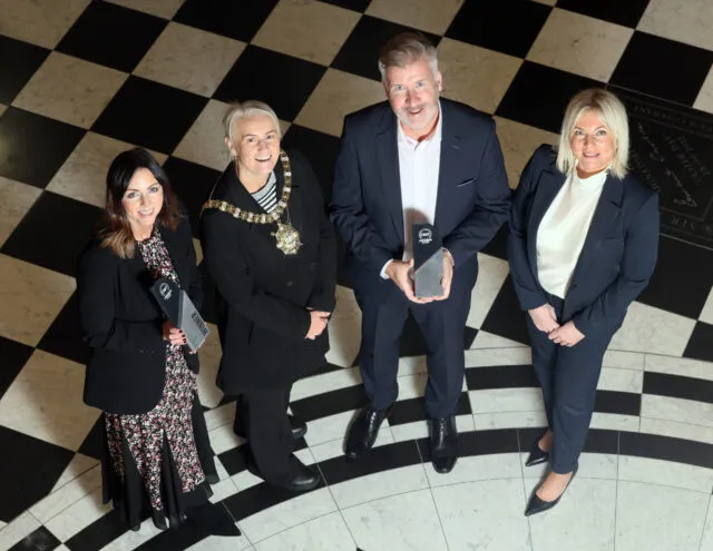 Group of 4 people standing on a black and white checked floor holding an award. One person is wearing the Lord Mayor Chain